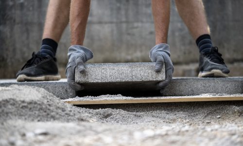 Low angle view of a man a worker wearing protective working gloves placing cement tile on a prepared surface while aving an outdoor patio. DIY project.