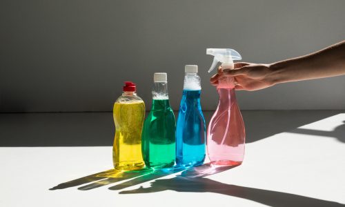 partial view of hand holding spray bottle and various cleaning fluids on white
