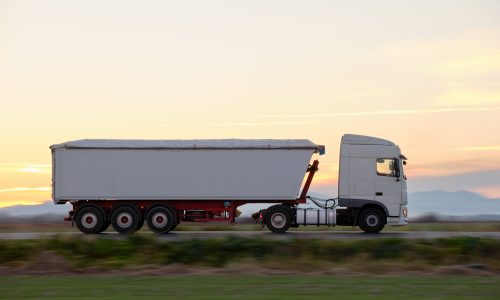 Semi-truck with tipping cargo trailer transporting sand from quarry driving on highway hauling goods in evening. Delivery transportation and logistics concept.